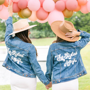 Two women in hats stand hand in hand wearing custom retro jean jackets.