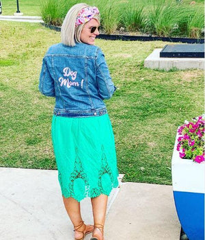 A woman stands outside showing off her custom jean jacket which reads "Dog Mom!"