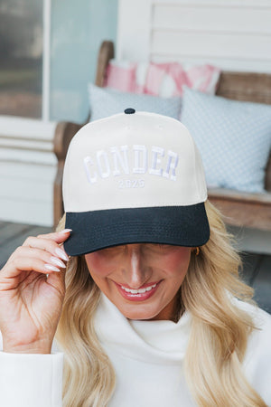 Close-up photo of a woman smiling and slightly tipping a baseball cap with her left hand. The cap features a cream crown and a navy blue bill, embroidered with 'CONDER 2025' in shimmering silver letters. She wears a white turtleneck sweatshirt and her long blonde hair is styled in waves, complementing her cheerful expression. The background reveals a cozy porch setting with cushioned benches, creating a warm and inviting atmosphere.