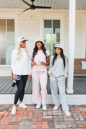 Photo of three women standing on a brick porch in front of a white house with wooden columns. From left to right: the first woman is in a white sweatshirt and black leggings, wearing a white cap with 'Forever' in black. The middle woman is dressed in a light pink set wearing a white cap with 'In My Mom Era' in black. The third woman is in a grey sweatshirt and pants, wearing a white cap with 'Oxford' in black. All three are smiling and casually posed, conveying a relaxed, friendly vibe.