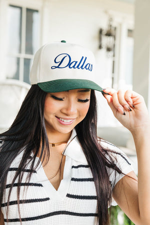 Image of a woman with long dark hair, smiling and adjusting the brim of her baseball cap with her left hand. The cap is cream-colored with a dark green bill, embroidered with the word 'Dallas' in curved navy script. She is wearing a white and black striped V-neck sweater. The background features a blurred porch setting of a house, adding a casual and friendly ambiance to the scene.