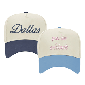 Image of two baseball caps with custom embroidery. The left cap has a cream crown and a navy blue bill, featuring the word 'Dallas' embroidered in elegant navy script. The right cap also has a cream crown but with a light blue bill, displaying the phrase 'spritz o'clock' in playful pink cursive. Both caps are structured with a classic design and have adjustable snaps at the back, suitable for personalized style or city pride.