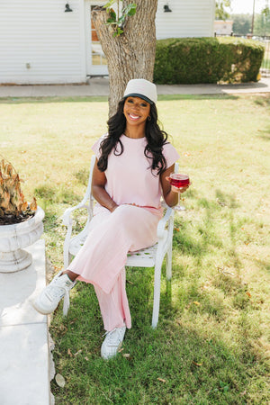 Photo of a smiling woman seated in a white garden chair, holding a cocktail. She is dressed in a light pink, soft cotton jumpsuit and wears a black cap with 'spritz o'clock' embroidered in pink cursive. The setting is a sunny backyard with a large tree and a white house in the background, creating a relaxed and cheerful atmosphere suitable for a casual outdoor gathering.