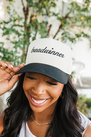 Image of a joyful woman adjusting a baseball cap with her right hand. The cap has a cream crown and a black bill, embroidered with the word 'breadwinner' in cursive black script. She is smiling broadly, showcasing her white tank top and delicate necklace, set against a blurred garden backdrop with lush greenery. Her expression reflects a playful and confident mood, emphasizing the empowering message on her cap.