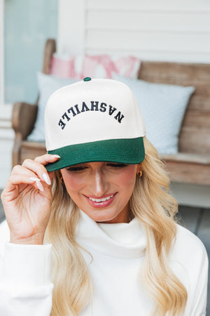 Close-up photo of a woman smiling and tipping her baseball cap with her left hand. She is wearing a cream and dark green cap with the word 'Nashville' embroidered in black letters across the front. Her attire includes a white turtleneck sweatshirt. The background features a cozy porch setting with a bench and cushions, adding a warm, homely feel to the scene.