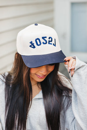 Image of a woman with long dark hair, smiling as she tips the brim of her baseball cap. The cap is cream-colored with a navy blue bill, and the word '75204' is embroidered in bold navy letters across the front. She wears a grey zip-up hoodie and stands in front of a blurred house with a blue door, giving the photo a casual, friendly vibe.