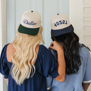 Image showing the backs of two women standing shoulder to shoulder, each wearing a baseball cap and blue tops. The woman on the left wears a cap with a cream crown and a dark green bill, embroidered with 'Dallas' in navy script. The woman on the right sports a cap with a cream crown and a navy bill, showing the word '75204' in navy, mirrored script. Both are set against a backdrop of a light blue wooden door, highlighting a casual, friendly moment.