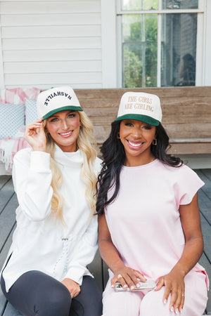 Photo of two women seated together on a porch bench, smiling at the camera. The woman on the left, with blonde hair, wears a white turtleneck sweatshirt and a cream cap with 'Nashville' in green letters. The woman on the right, with black hair, wears a light pink blouse and a cream cap that reads 'THE GIRLS ARE GIRLING' in green letters. The background features a wooden porch with large windows, enhancing the warm and friendly atmosphere of the setting.