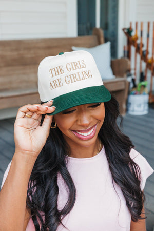 Photo of a joyful woman tipping her baseball cap with her right hand. The cap features a cream upper and a dark green bill, with the phrase 'THE GIRLS ARE GIRLING' embroidered in gold.