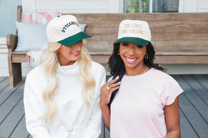 Photo of two women smiling and sitting close together on a wooden porch bench. The woman on the left, with long blonde hair, wears a white and dark green cap embroidered with 'Nashville' in black letters, paired with a white turtleneck sweatshirt. The woman on the right, with long black hair, wears a cream and dark green cap stating 'THE GIRLS ARE GIRLING' in gold letters, paired with a light pink blouse. The background features a cozy porch setting with pillows and a wooden railing.