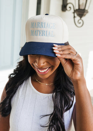 Image of a smiling woman adjusting a baseball cap with her left hand. The cap has a cream crown and a navy blue bill, embroidered with the phrase 'MARRIAGE MATERIAL' in light blue thread. The woman is wearing a white sleeveless top and a delicate necklace, standing in front of a blurred light-colored doorway background.