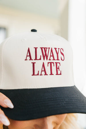 Close-up image of a baseball cap held by a person, visible only from the nose down. The cap has a white crown and a black bill, embroidered with the phrase 'ALWAYS LATE' in bold maroon letters.