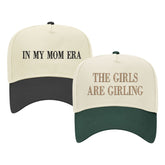 Image of two baseball caps. The left cap has a cream top with a black bill and features the phrase 'IN MY MOM ERA' embroidered in black. The right cap has a cream top with a dark green bill and displays the embroidered phrase 'THE GIRLS ARE GIRLING' in gold