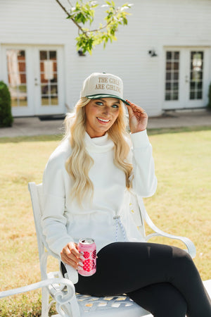 Photo of a woman sitting on a white garden chair outdoors, smiling and tipping her hat. She is wearing a cream cap with the phrase 'THE GIRLS ARE GIRLING' in gold letters, paired with a white turtleneck sweatshirt and black leggings.