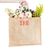 A person holds a pink and orange monogrammed jute bag filled with flowers.