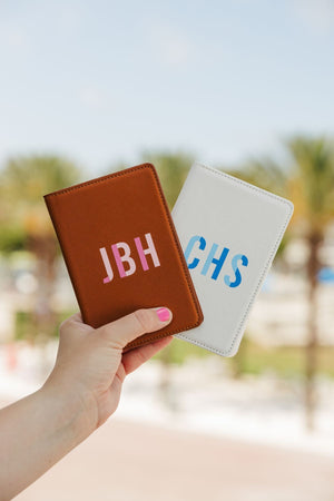 A person holds up a tan passport holder with a pink monogram and a white passport holder with a blue monogram.