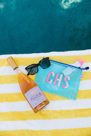 A blue pool bag is customized with a pink monogram and placed at the pool with a bottle of wine and some sunglasses.