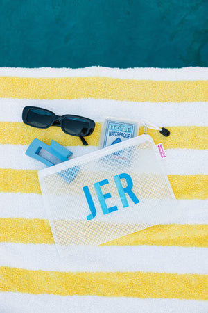 A white pool bag with a blue monogram is placed by the pool with some sunglasses and playing cards.