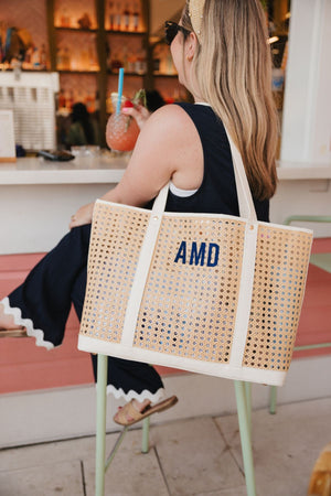 A girl sits at a bar wearing a cane tote that is customized with a navy embroidered monogram.