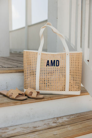 A cane tote with a navy embroidered monogram is placed on a dock next to a pair of sandals.