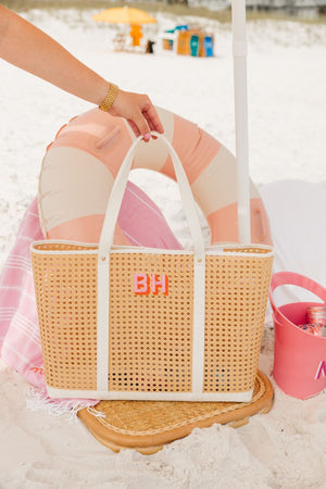 A cane tote is placed on a beach chair and is customized with a pink and orange monogram.