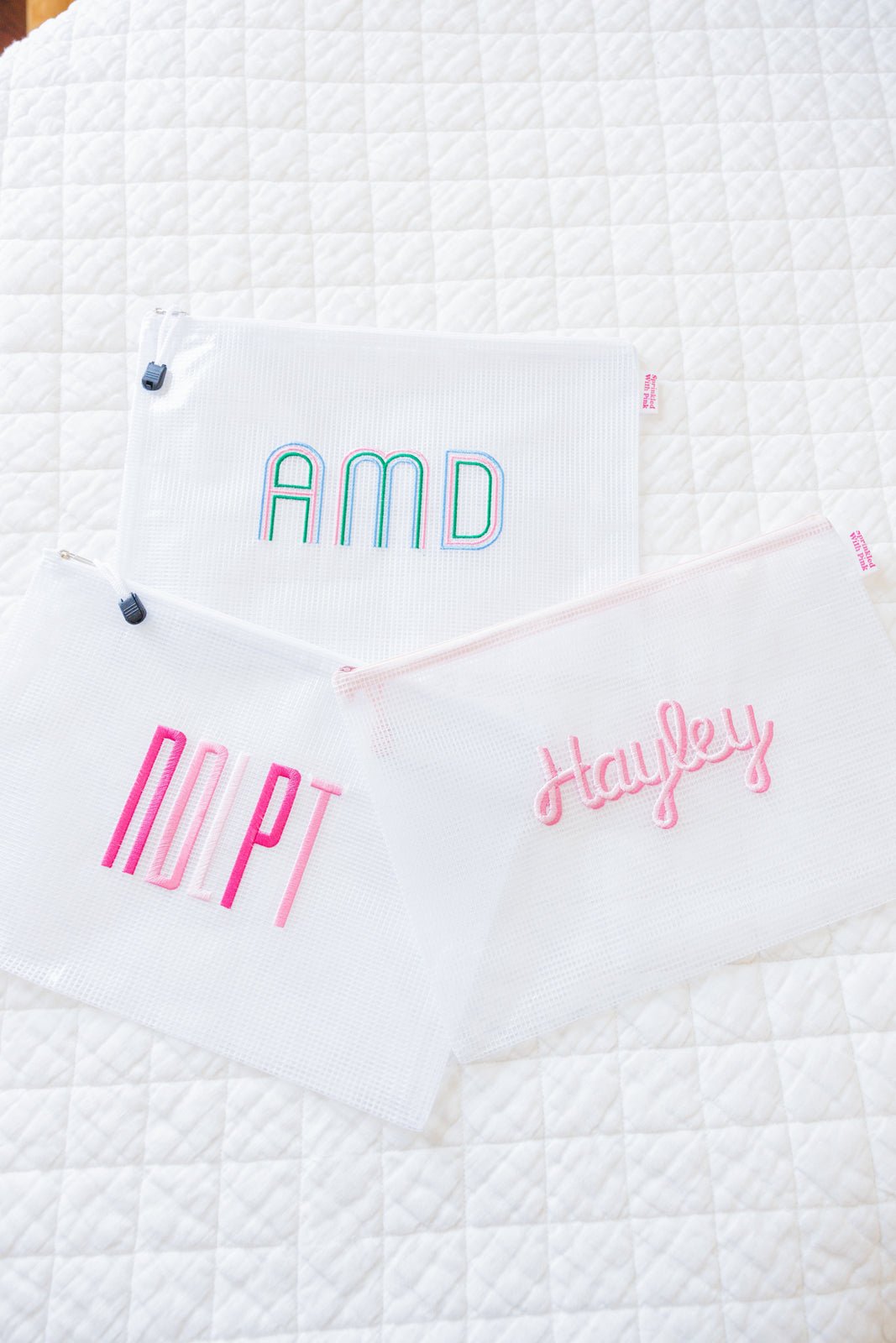 Three custom embroidered mesh pouches displayed against a white background. The first pouch is pink with the name 'Harper' in script embroidery, the second pouch is white with the monogram 'EMG' in pink embroidery, and the third pouch is blue with the name 'Sophia' in block embroidery.