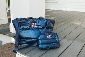 A set of navy blue quilted bags with personalized red 'KL' monograms on a wooden deck. The larger duffel bag and a smaller cosmetic bag are designed for both style and functionality, suited for travel or daily use. The setting includes architectural elements of a white porch, capturing a serene outdoor vibe that complements the stylish utility of the bags.