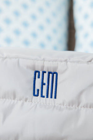 Close-up of a taupe quilted bag with a personalized monogram 'CEM' in vibrant blue stitching. The detailed view highlights the precise embroidery and texture of the fabric, set against a backdrop of soft blue patterned cushions, adding a touch of elegance and personal style to the item.