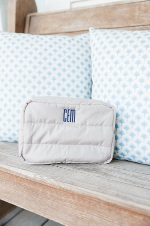 A light beige quilted travel bag featuring a personalized 'CEM' monogram in blue, positioned on a wooden bench. Behind the bag, two matching cushions with a blue and white floral pattern add a refreshing and decorative touch to the scene, emphasizing a tranquil and stylish setting.