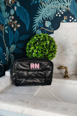 A stylish black quilted cosmetic bag with a vibrant pink 'RN' monogram positioned on a marble bathroom countertop. The elegant setting is enhanced by a detailed botanical wallpaper in blue and green hues and a decorative green topiary, adding a touch of luxury and personalization to the modern bathroom decor.