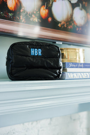 A black quilted cosmetic bag with a custom 'HBR' monogram in light blue displayed on a mantelpiece, set against a seasonal backdrop featuring a framed picture of pumpkins. The elegant setting includes a silver candle jar labeled 'McKinney' and enhances the personal charm and seasonal decor of the home.