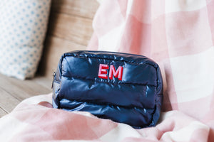 A cozy setting featuring a navy quilted cosmetic bag with a bold 'EM' monogram in red, resting on a soft pink and white checkered blanket. The backdrop includes a rustic wooden surface and a cushion with a blue pattern, enhancing the homey and personalized feel of the scene.