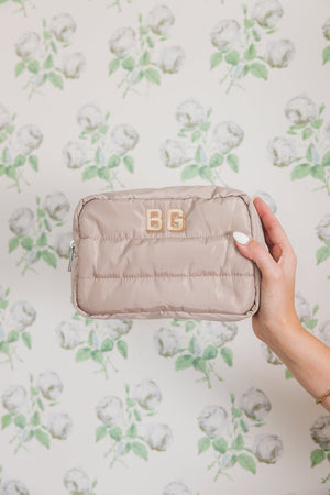 A taupe quilted cosmetic bag with a golden 'BG' monogram held up against a wallpaper adorned with a delicate pattern of green and white roses. The image highlights the elegance and personalized touch of the bag, showcasing its practical size and stylish design against a floral backdrop that adds a romantic aesthetic.