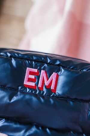 Close-up of a navy blue quilted bag featuring a bold 'EM' monogram in vibrant pink. The detail shot highlights the intricate stitching and texture of the embroidery, set against a soft pink background that subtly complements the monogram's color, adding a personalized and stylish element to the accessory.