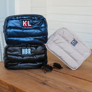 Three quilted travel bags displayed on a wooden surface, showcasing personalized monogram embroidery. The stacked bags in the foreground are black; the top one features 'KL' in bold red, and the middle one has 'HBR' in light blue. The third bag, standing alone to the right, is white with 'LH' in blue. A pair of sunglasses lies beside the bags, adding to the travel-ready theme.