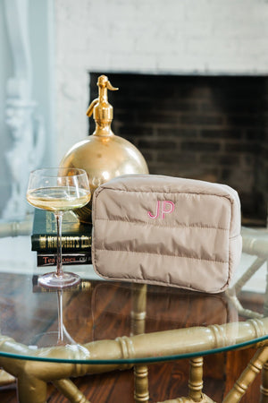 An elegant taupe cosmetic bag with a pink 'JP' monogram positioned on a glass coffee table amidst luxurious decor. Accompanying the bag is a glass of white wine, vintage books, and a golden siphon, set against a sophisticated backdrop of a dark fireplace and ornate white molding. The setting reflects a refined and personalized lifestyle, highlighting the bag’s chic design and custom touch.