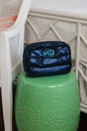 A navy blue quilted cosmetic bag with a turquoise 'MD' monogram rests atop a vibrant green ceramic stool. The setting features intricate patterns on the stool, complementing the sleek simplicity of the bag. Positioned beside a white wicker chair with a peek of orange fabric, the scene merges practicality with a pop of color and personal flair.