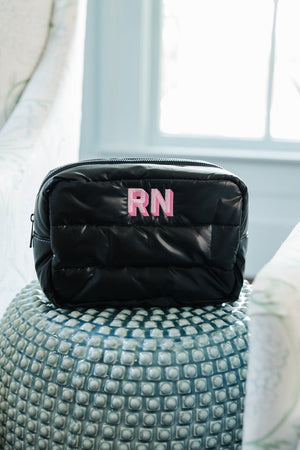 A black quilted cosmetic bag with a vibrant pink 'RN' monogram sits atop a textured silver ottoman. The bag is strategically placed to catch the soft natural light from a nearby window, enhancing its sleek design and personalized detail in a stylish home setting.