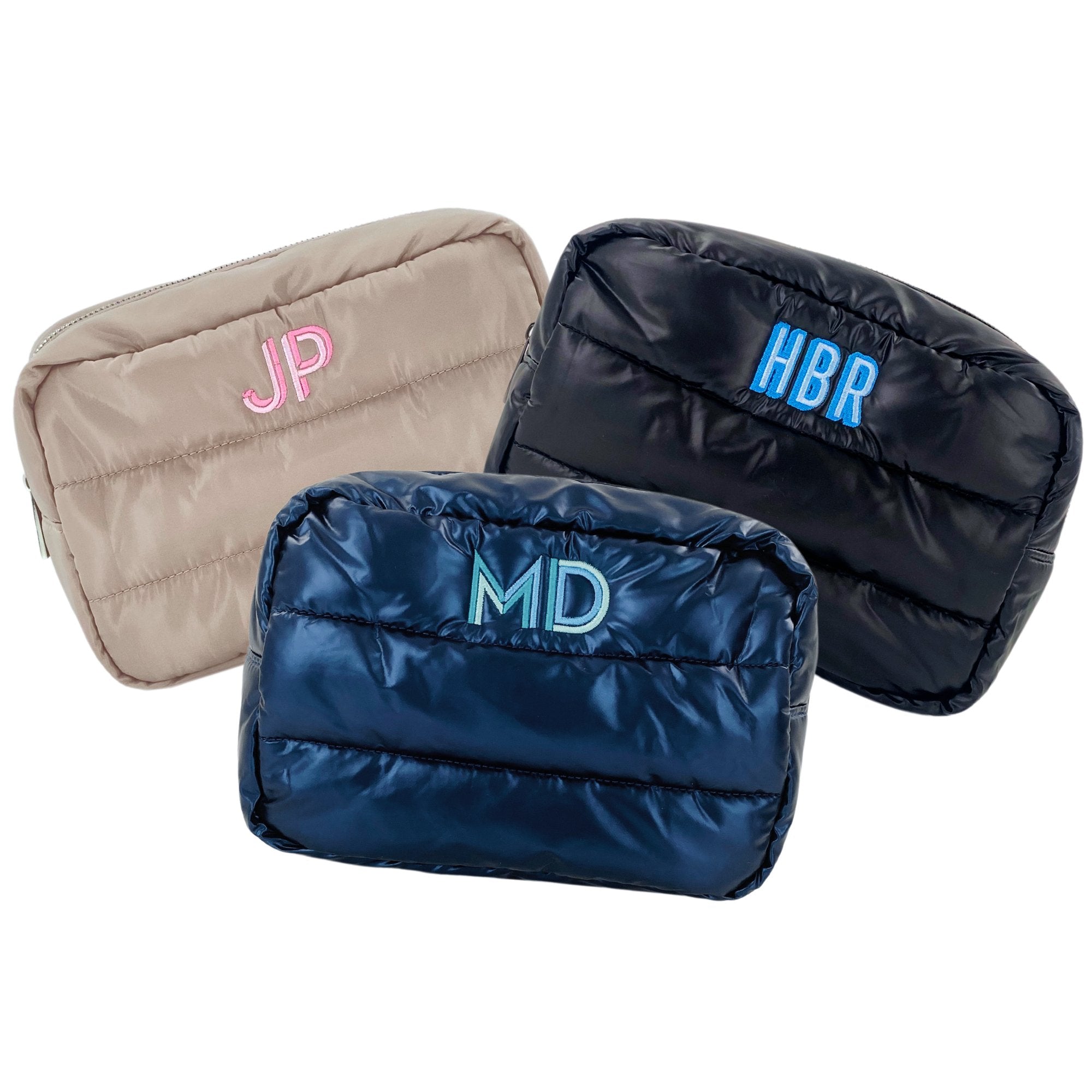 Three custom embroidered puffer pouches displayed against a white background, featuring a beige pouch with pink 'JP' monogram, a black pouch with blue 'HBR' monogram, and a navy pouch with teal 'MD' monogram.