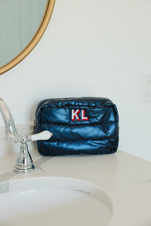 A navy blue quilted cosmetic bag with a vibrant red 'KL' monogram sits on a white marble countertop next to a silver faucet. Behind the bag, a round gold mirror adds a touch of elegance to the minimalist bathroom setting, highlighting the bag's sleek design and personalized detail.