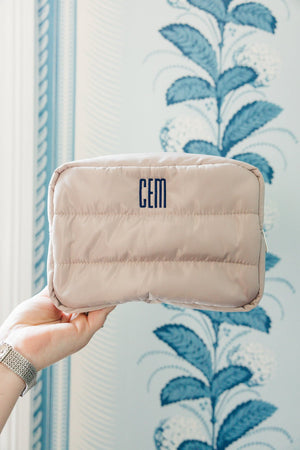 A hand holds a taupe quilted cosmetic bag featuring a personalized 'CEM' monogram in bold blue, against a backdrop of elegant blue and white floral wallpaper. The image captures the sophisticated stitching and texture of the bag, highlighting its custom design and the harmonious blend with the decorative setting.