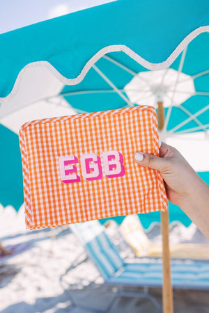 A person holds up an orange pouch with a pink monogram in front of a blue umbrella.