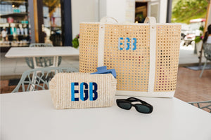 A straw pouch and a cane tote are customized with a blue monogram and placed on a table with some sunglasses.