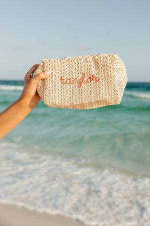 A person holds up a straw pouch with an orange embroidered name on it.