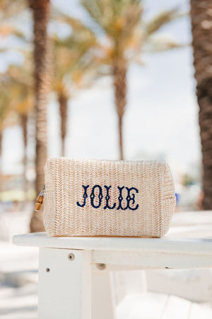 A straw pouch with a name embroidered in navy thread is placed on a beach chair