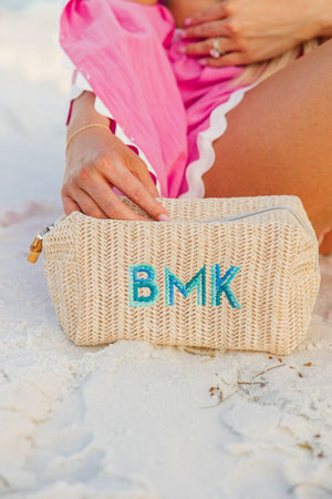 A straw pouch is placed in the sand at a beach and us customized with a blue monogram.