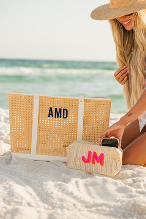 A girl on the beach puts her phone into a straw pouch with a pink and orange monogram embroidered on it.