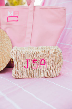 A straw pouch is customized with a pink monogram and placed on a pink towel next to a pink coated canvas tote.