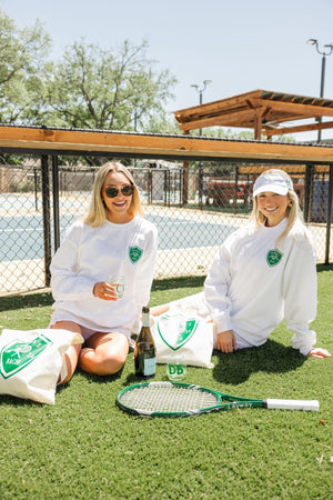 Two girls on a tennis court wear matching white sweatshirts with green Last Swing Before The Ring sweatshirts