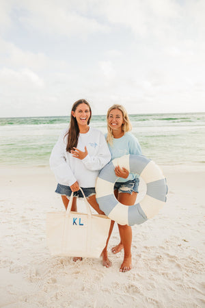 Two girls at the beach smile and pose with their custom bachelorette gear including a sweatshirt and a coated canvas tote. 